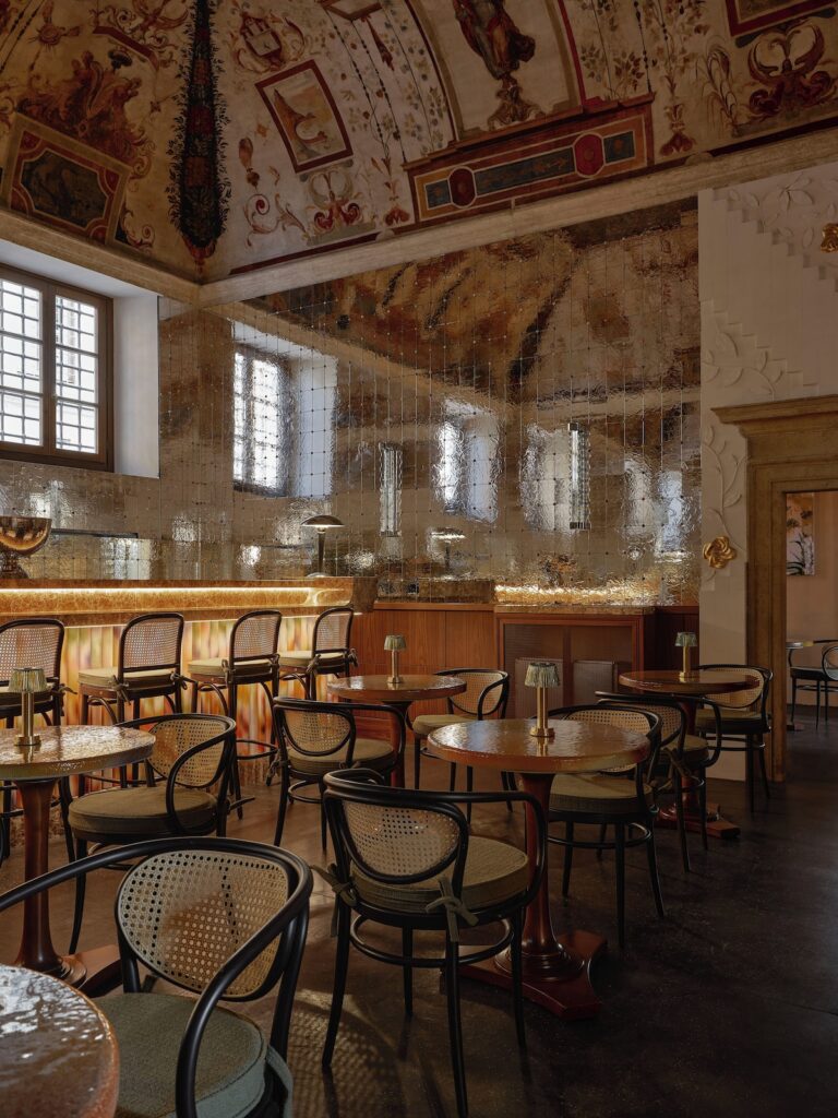 Bar do hotel Palazzo Talìa em Roma com afrescos históricos no teto, parede espelhada ondulada e mesas bistrô de madeira.