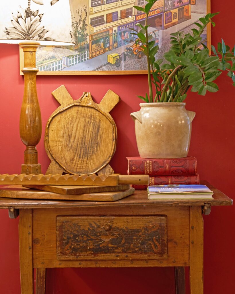 Pátina e palimpsesto na arquitetura: mesa rústica com pátina, objetos vintage, planta em vaso cerâmico e arte retrô em parede vermelha, estilo boho-chique. Arnaldo Danemberg Collection