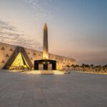 Fachada monumental em alabastro transl&uacute;cido e pra&ccedil;a com obelisco suspenso no Grand Egyptian Museum em Giz&eacute;.