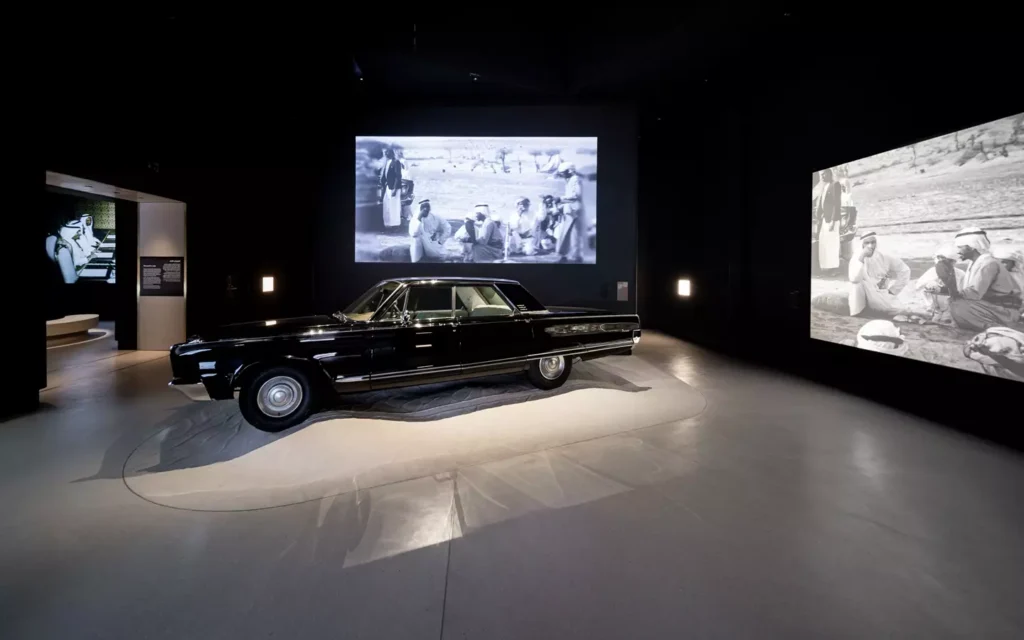 Galeria imersiva no Zayed National Museum com automóvel histórico clássico e projeções de imagens de arquivo em paredes escuras.