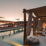 Cabanas de madeira &agrave; beira da piscina ao p&ocirc;r do sol no Branco Mykonos, cen&aacute;rio sofisticado de onde ficar em Mykonos.