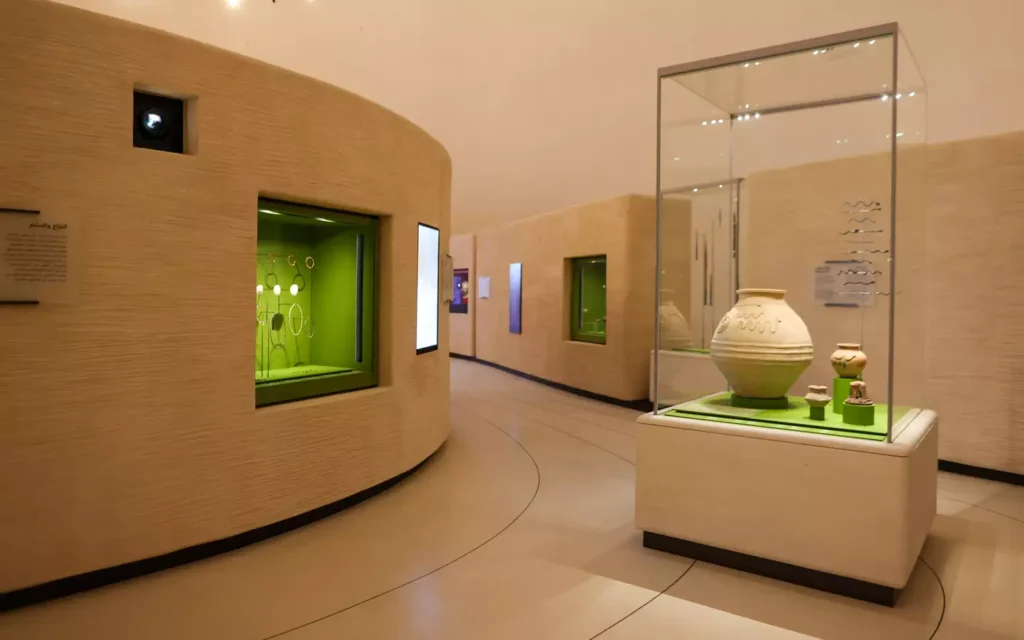 Corredor expositivo no Zayed National Museum com paredes curvas de textura arenosa e vitrines com cerâmicas e artefatos.