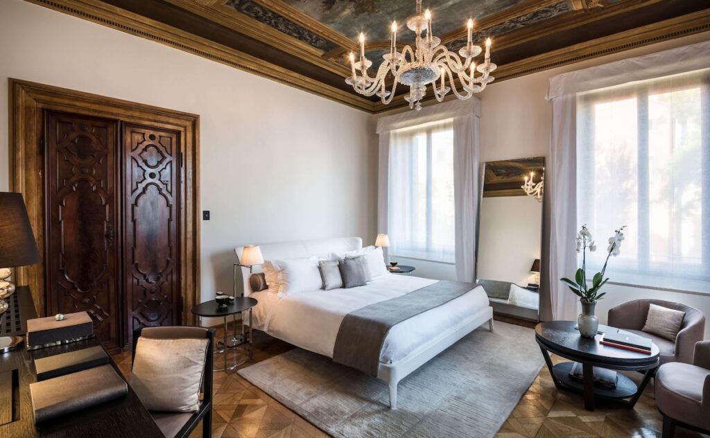 Quarto palaciano do Aman Venice com porta entalhada, afresco e lustre de Murano, requinte nos hotéis de Veneza.