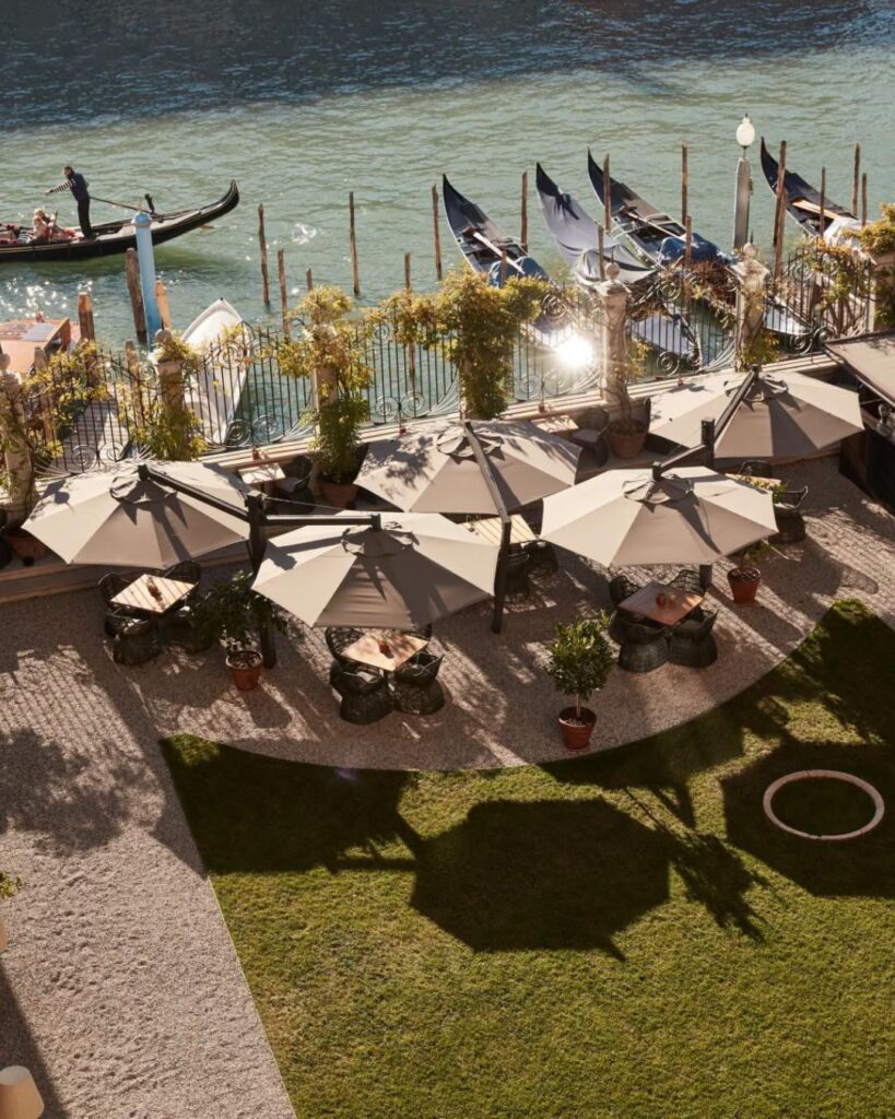 Jardim privativo à beira do Grand Canal no Aman Venice com mesas e ombrelones, exclusividade nos hotéis de Veneza.