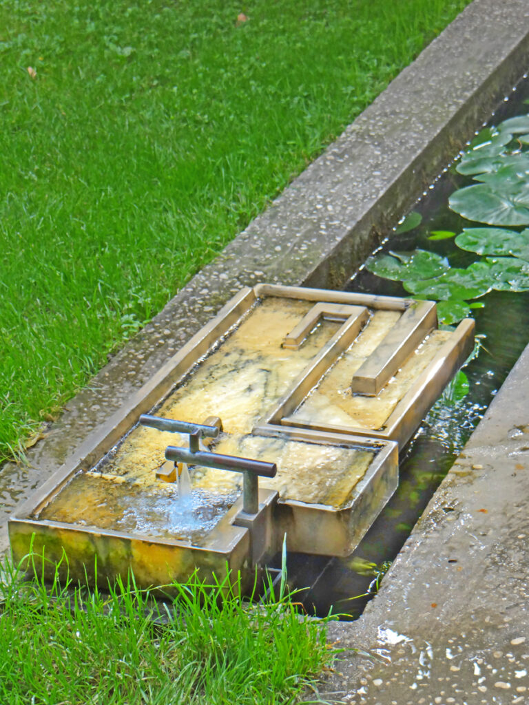 Dispositivo labiríntico em bronze e mármore da fonte no jardim da Fondazione Querini Stampalia com calha de ninfeias e relvado verde.