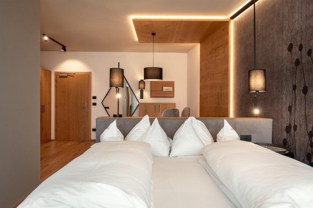 Quarto de casal com marcenaria iluminada e cabeceira estofada no Hotel Hubertus, sinônimo de acolhimento em hotéis de luxo nos Alpes italianos.