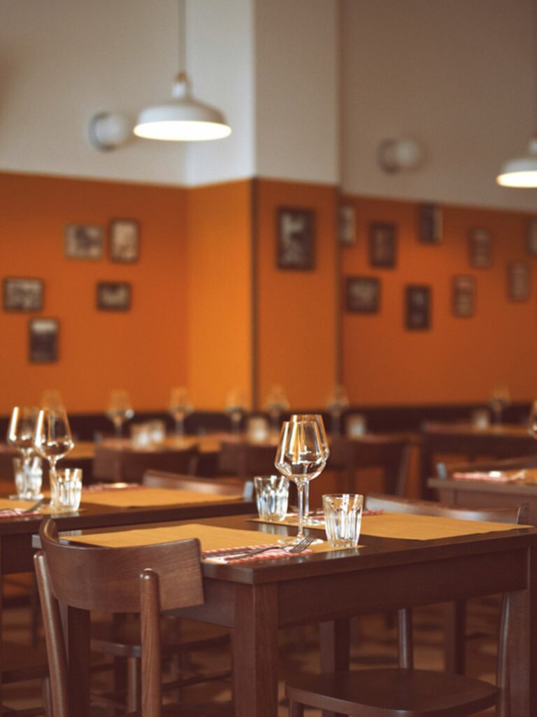Salão do restaurante Trippa Milano com mesas de madeira escura, cadeiras clássicas, paredes ocre e quadros vintage dos anos 50.