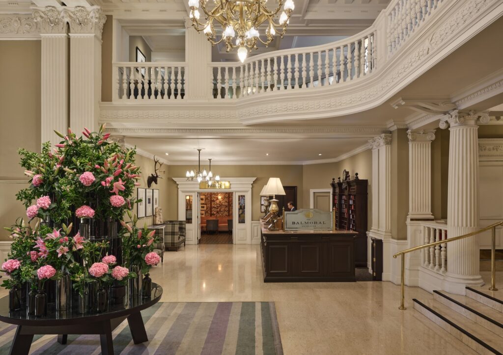 Lobby clássico do Balmoral hotel em Edimburgo com colunas ornamentadas, mezanino curvo, imponente lustre e exuberante arranjo floral sobre mármore.