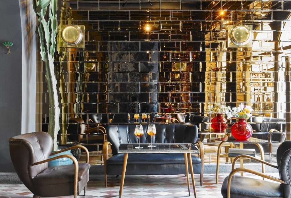 Gallery Bar no G-Rough Roma com revestimento de espelho âmbar, sofá capitonê vintage e piso geométrico.