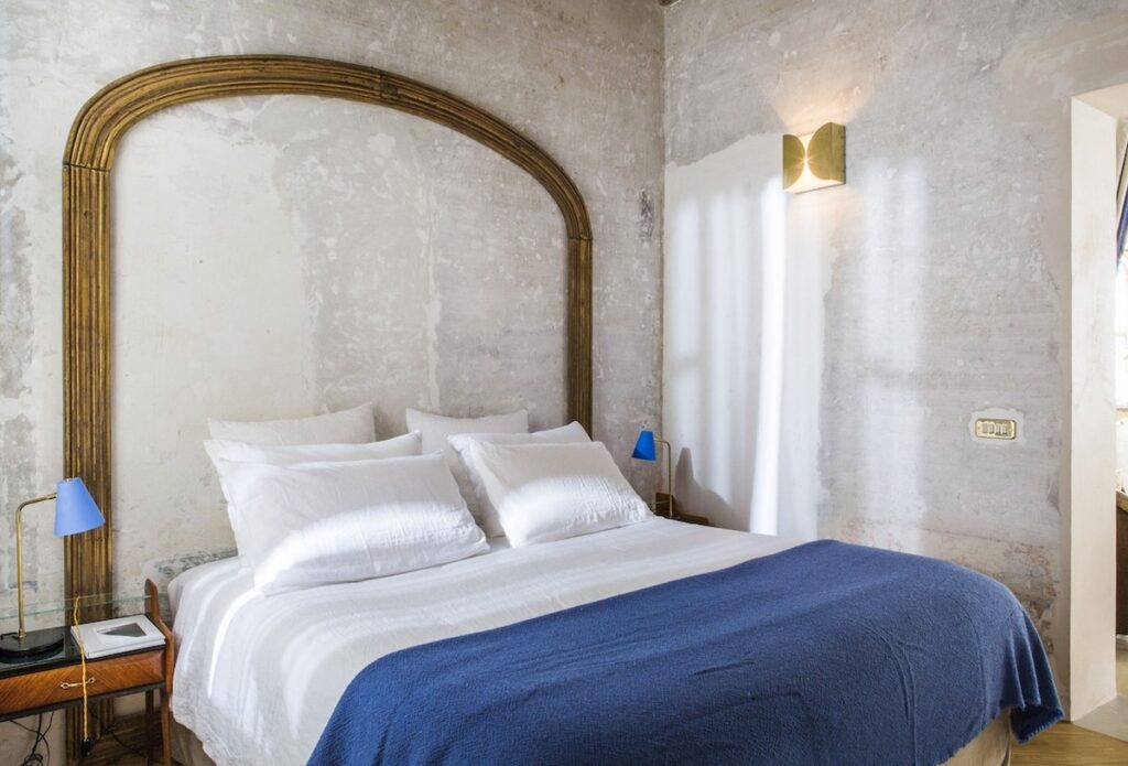 Detalhe da cama e arandela de latão em parede de textura rústica no hotel G-Rough Roma.
