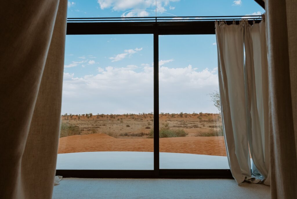 Esquadria piso-teto com cortinas de linho enquadrando as dunas de areia vermelha a partir de retiro no deserto.