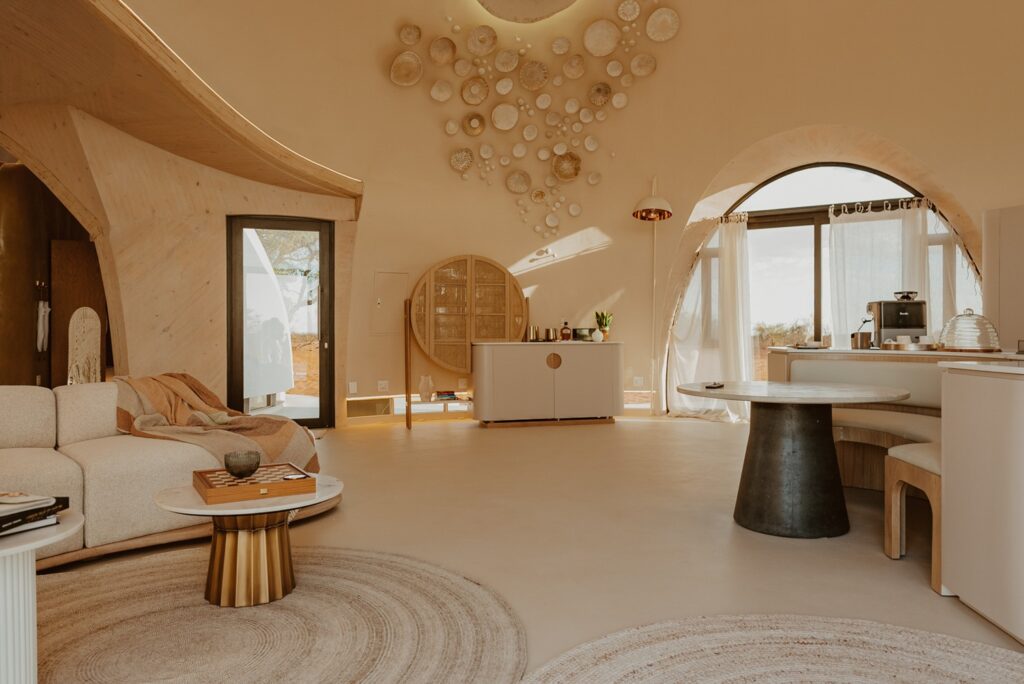 Living e cozinha integrada sob cúpula orgânica com mobiliário curvilíneo e luz natural no Reverie Kalahari Pod.
