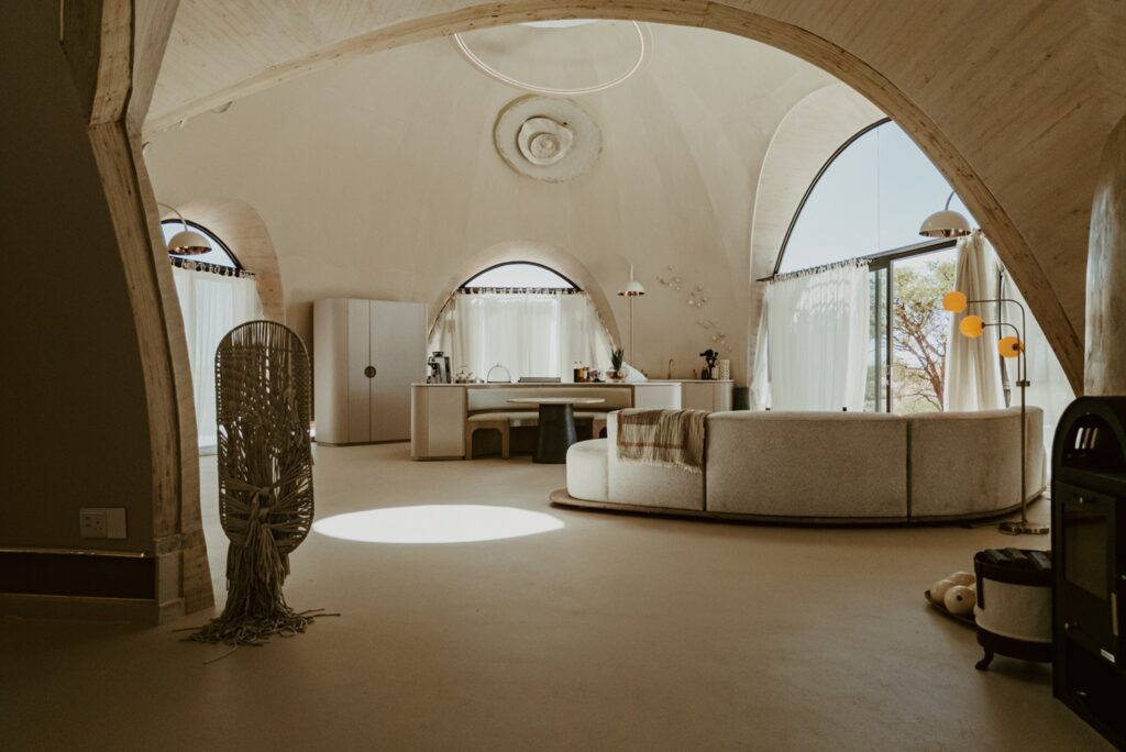 Living com sofá curvilíneo sob feixe de luz solar zenital projetado no piso monolítico do Reverie Kalahari Pod.
