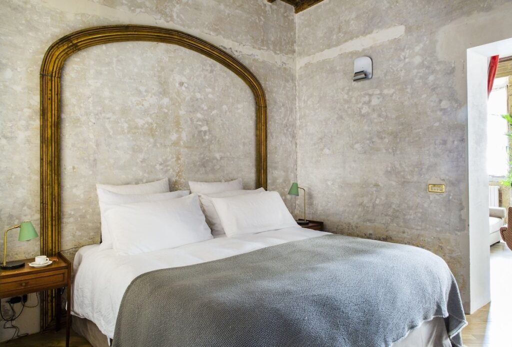 Dormitório da Suíte Pasquino no G-Rough Roma com cabeceira em arco dourado e roupa de cama de linho.