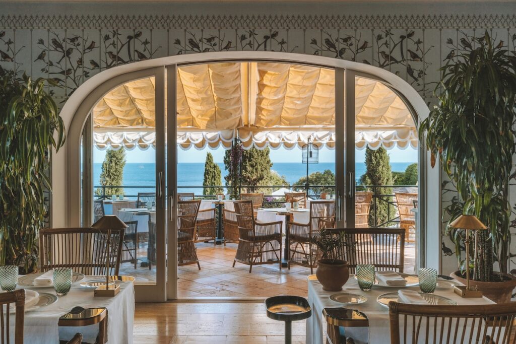 Salão com vista para o mar no Il Pellicano, destaque entre os restaurantes mais belos da Itália com cadeiras de vime e papel de parede.