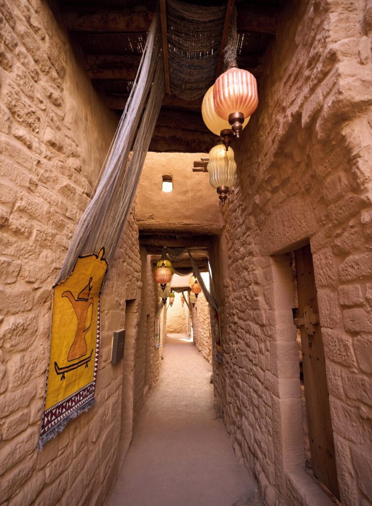 Corredor histórico entre paredes de pedra e luminárias de vidro em hotel no deserto de AlUla.