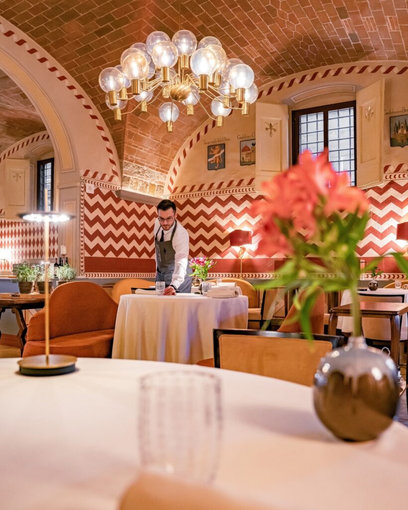 Salão sob abóbada de tijolos da Osteria di Passignano, com afrescos geométricos, lustre de design em latão e mesas redondas de linho.