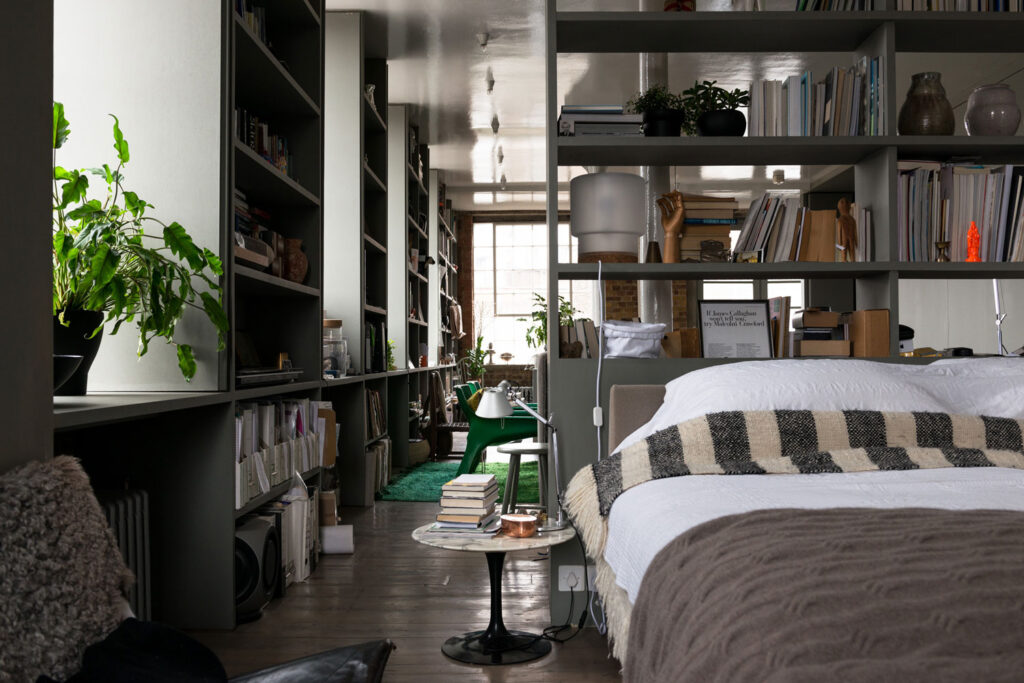 Quarto integrado de loft assinado por Ilse Crawford com estante modular cinza e cama com texturas acolhedoras.