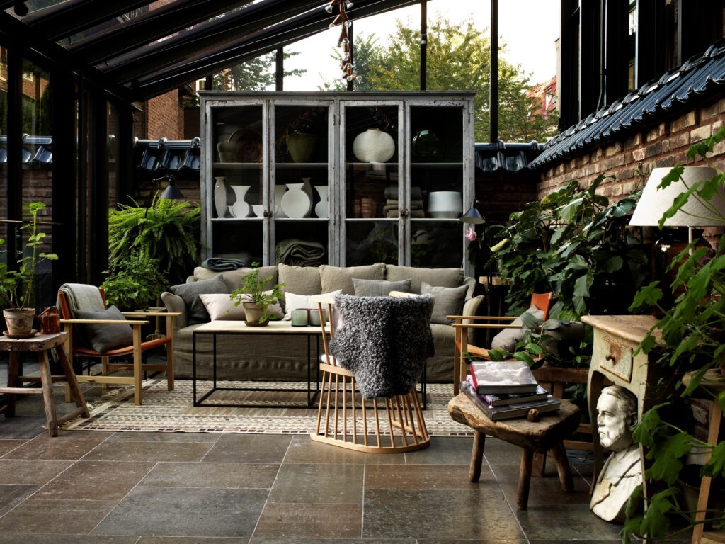 Jardim de inverno envidraçado do Ett Hem por Ilse Crawford com cristaleira vintage, piso de pedra e vegetação.