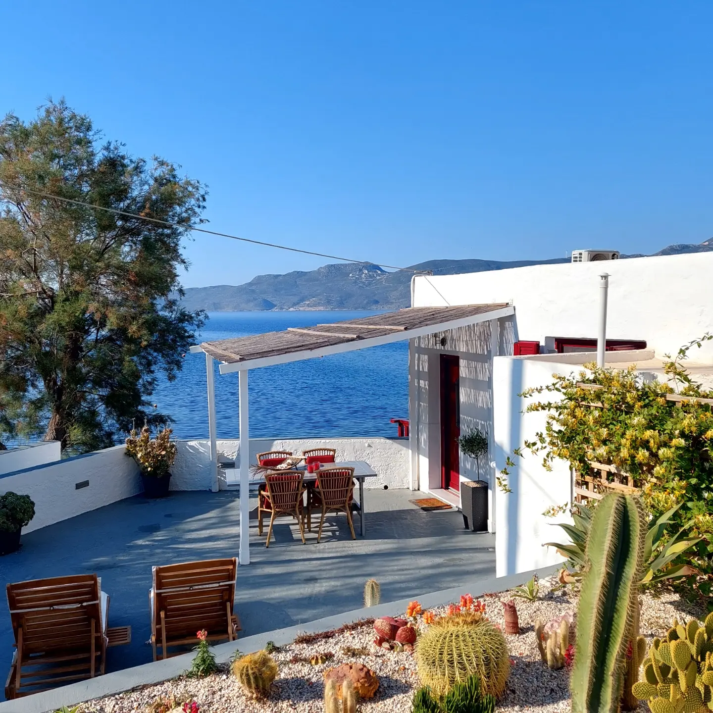 Terraço panorâmico de Syrmatas na costa grega com pérgula ripada, piso cimentício, jardim xerófilo de cactos e vista para o Mar Egeu.