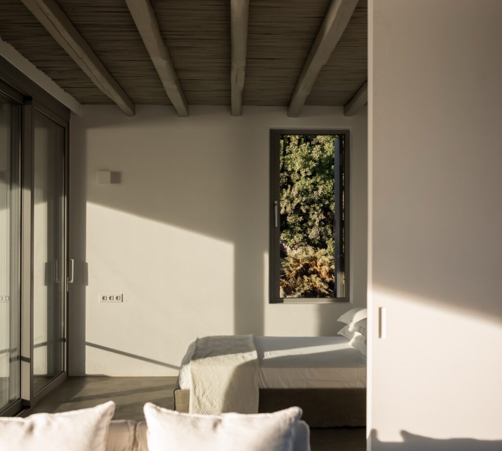 Suíte minimalista no Skinopi Lodge em Milos com vigas rústicas, piso em microcimento, janela panorâmica e diálogo com Syrmatas.
