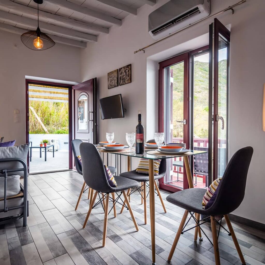 Área de refeições em Syrmatas convertidas em refúgio boutique, com teto de vigas caiadas, caixilhos bordô e mesa contemporânea com vista.