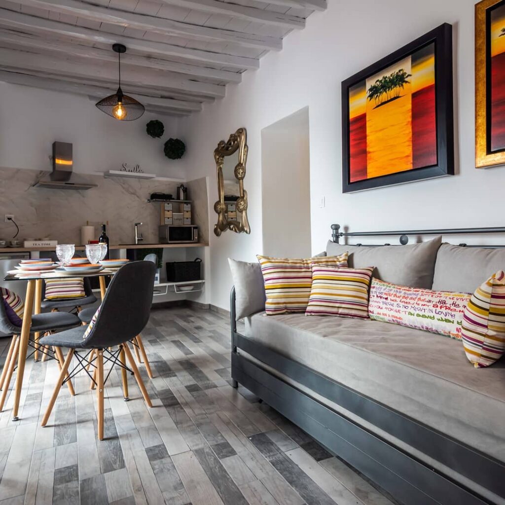 Living integrado de Syrmatas em Milos com daybed tubular, cozinha compacta em mármore, espelho rococó e teto de madeira rústica caiada.
