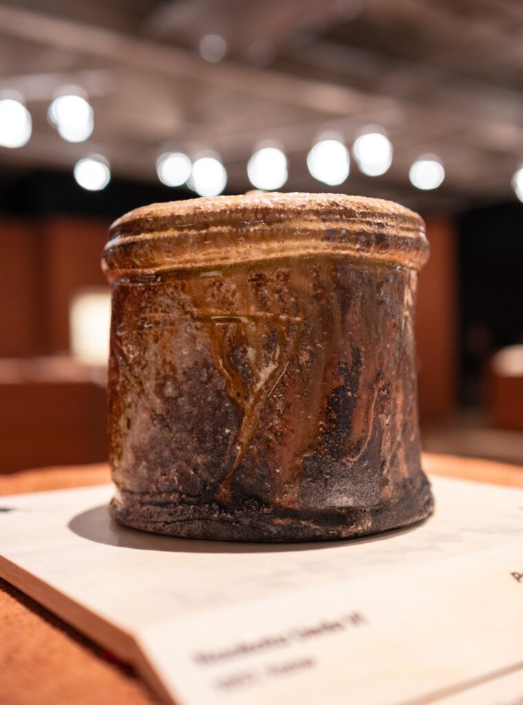 Recipiente cerâmico rústico com esmalte escorrido sobre base de madeira clara em exposições em São Paulo na Japan House.