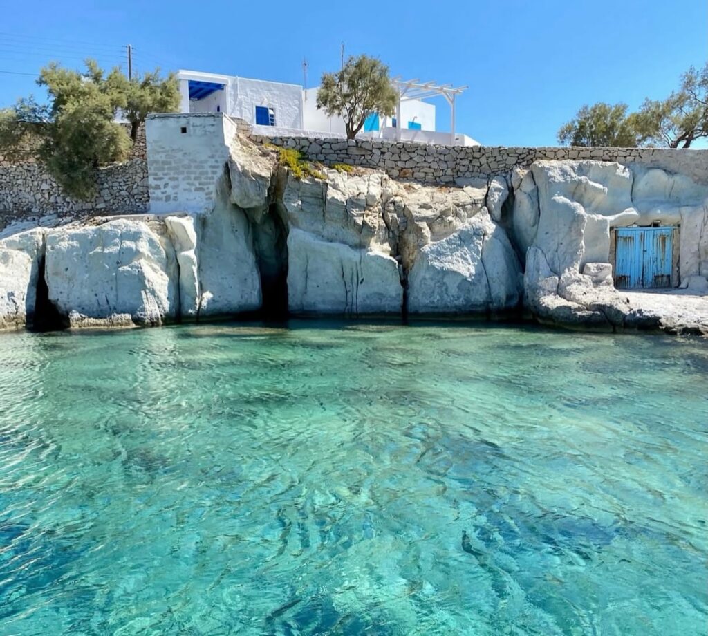 Enseada de águas cristalinas em Milos com Syrmatas escavadas na rocha vulcânica e a arquitetura do Skinopi Lodge na encosta superior.
