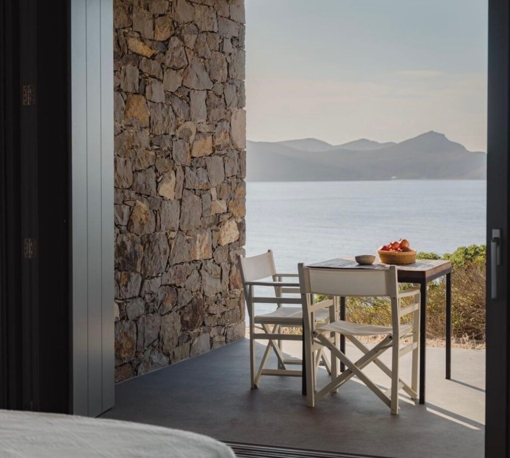 Varanda privativa do Skinopi Lodge com parede de pedra vulcânica, mesa bistrô e vista para o mar de Milos e suas Syrmatas.