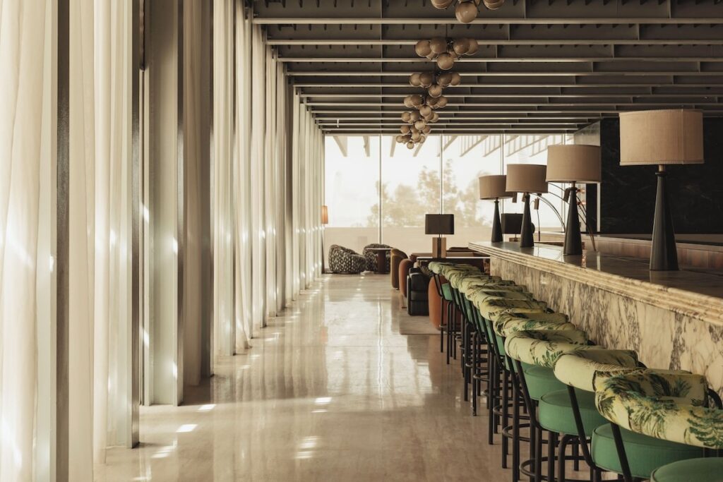 Balcão de bar em mármore com banquetas botânicas e iluminação filtrada por cortinas no hotel Ned Doha.