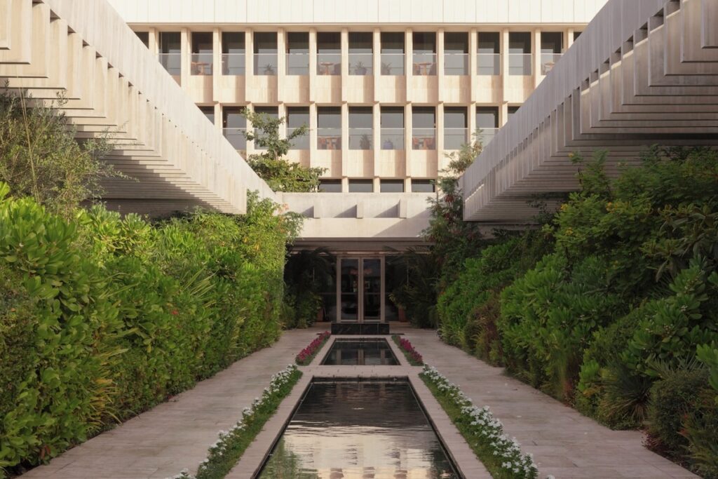 Pátio ajardinado com espelho d'água linear e pérgolas de concreto simétricas na arquitetura do Ned Doha.