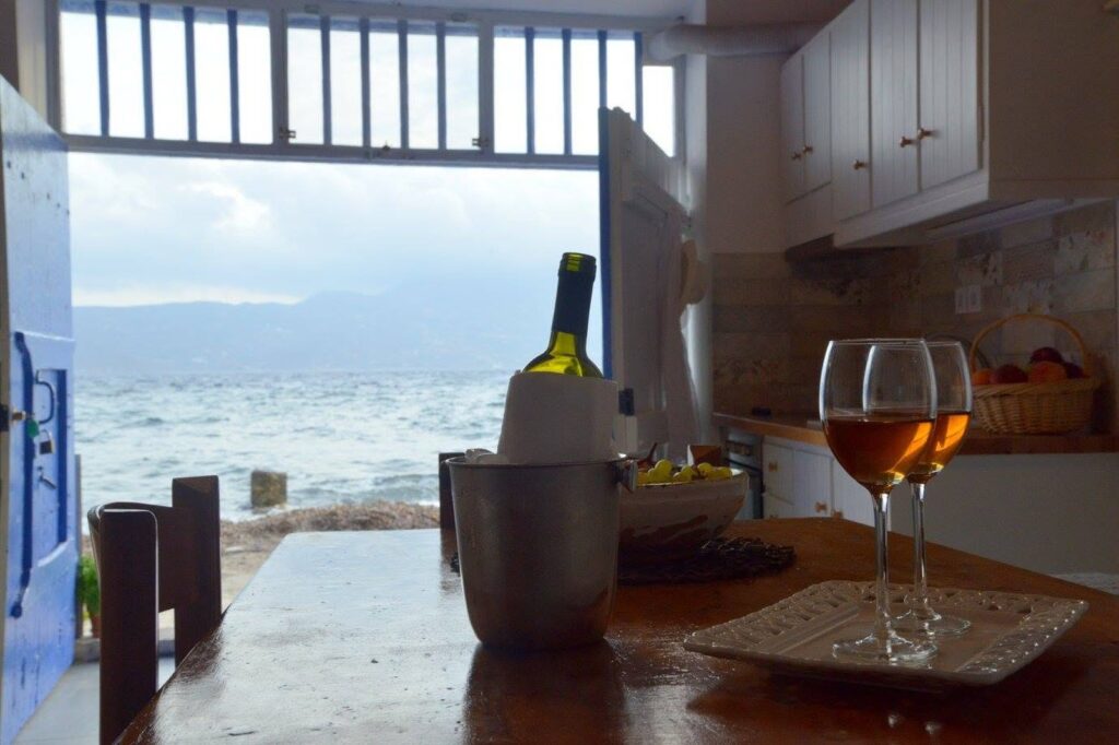 Cozinha e sala térrea de Syrmatas na Blue Sea House com portal azul cobalto aberto rente ao mar e mesa de madeira rústica.