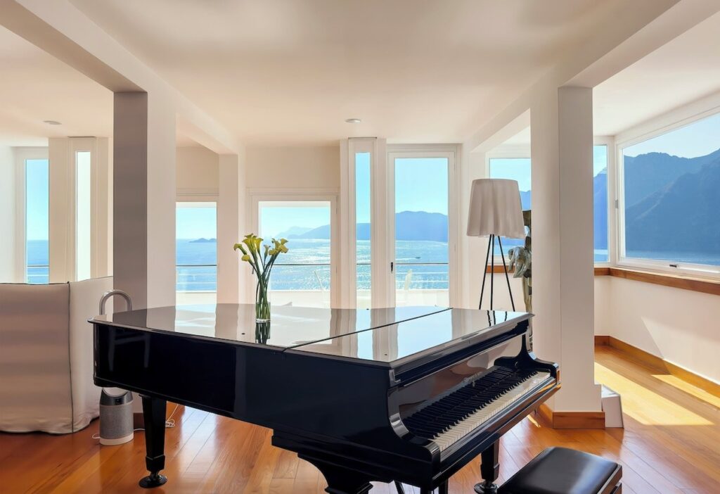 Salão de estar com piano de cauda preto e amplas janelas para o mar no hotel Casa Angelina.
