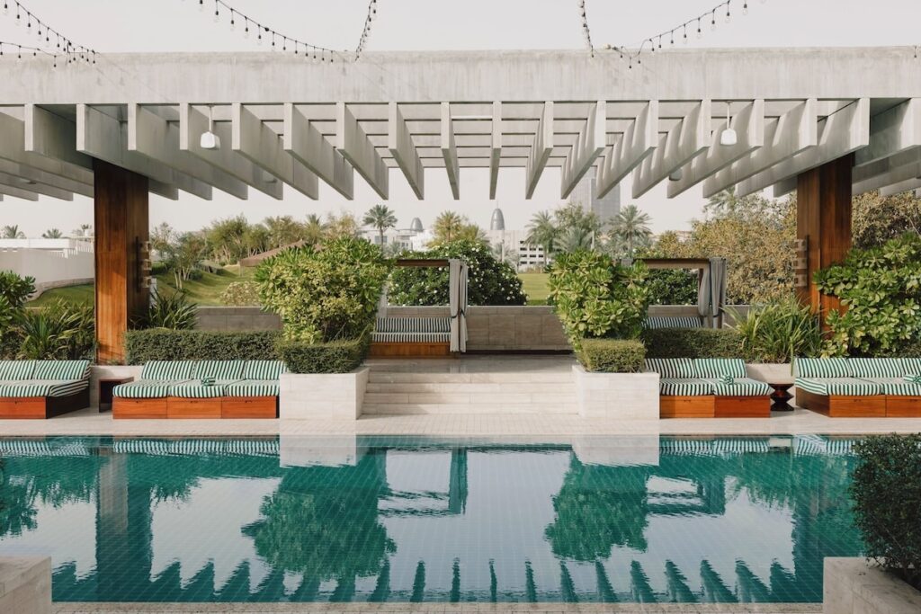 Piscina turquesa com espreguiçadeiras listradas e pérgola de concreto aparente no hotel Ned Doha.