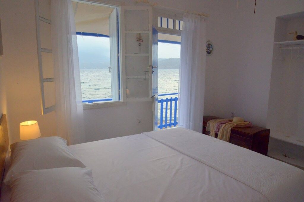 Dormitório minimalista em Syrmatas na Blue Sea House com enxoval branco, nichos em alvenaria caiada e vista para as águas de Milos.