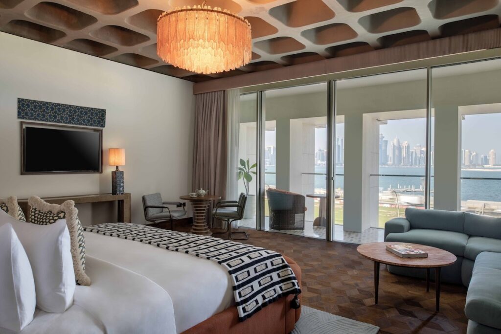 Suíte com teto de concreto aparente, lustre de Murano e varanda com vista para o mar no Ned Doha.