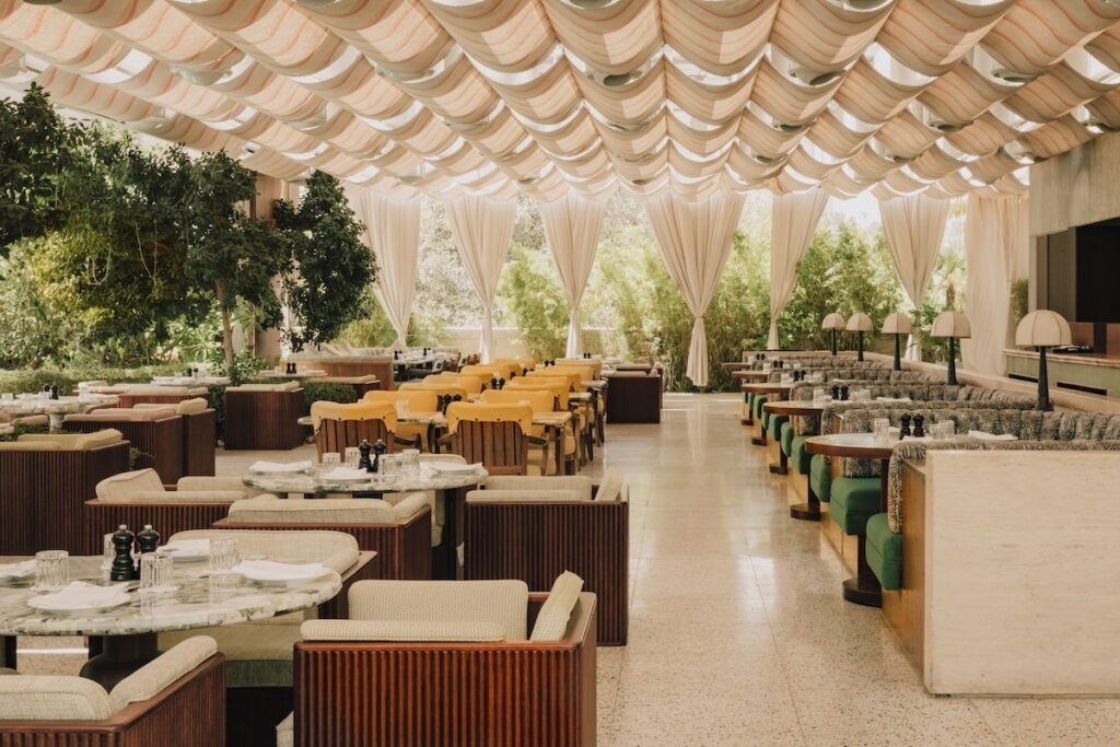 Salão de restaurante com teto em drapeados têxteis, booths em veludo verde e mesas de mármore no Ned Doha.